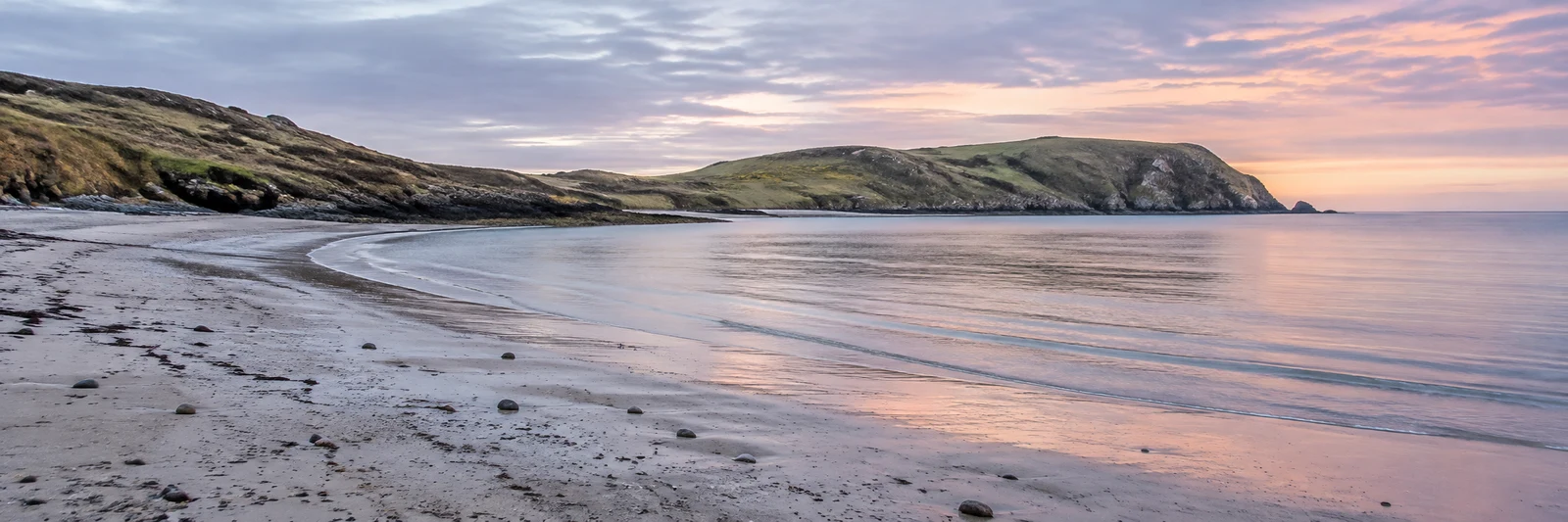 An empty crescent bay with wet, pebble-strewn sand in the foreground, calm water, and grassy rocky headlands beneath layered pink and violet clouds.