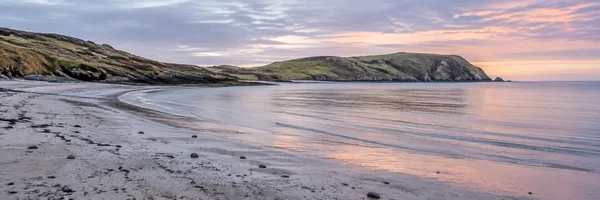An empty crescent bay with wet, pebble-strewn sand in the foreground, calm water, and grassy rocky headlands beneath layered pink and violet clouds.