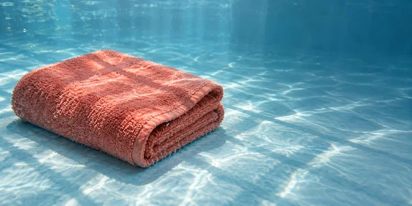 A neatly folded coral terry towel rests on a pale blue underwater floor, with bright rippling light patterns across the fabric and surrounding water.