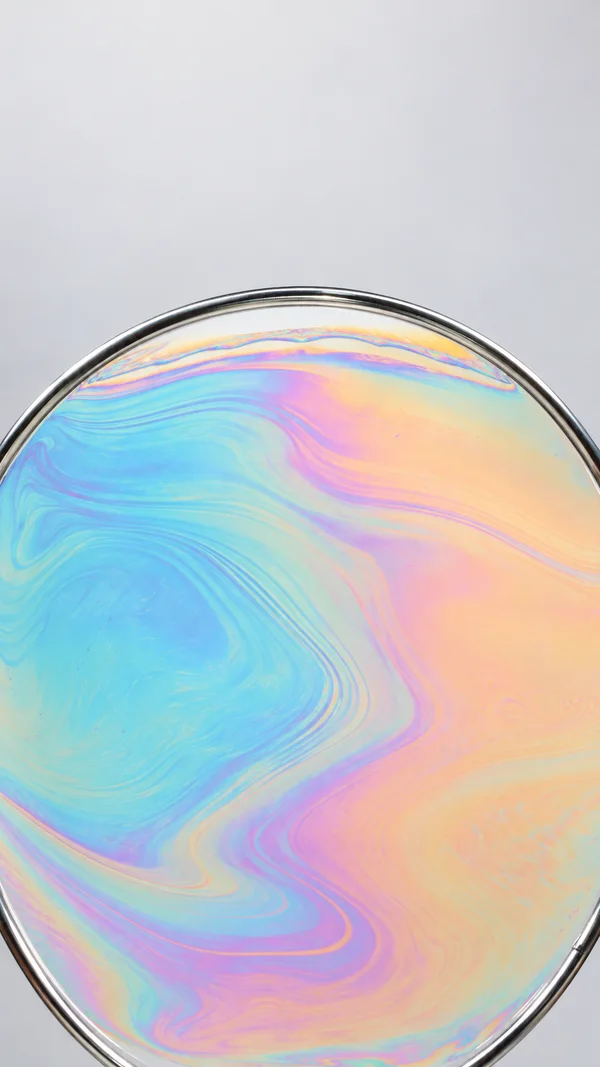 A close-up of a large iridescent film stretched across a polished chrome loop, with flowing cyan, peach, and lilac bands against a pale gray background and…