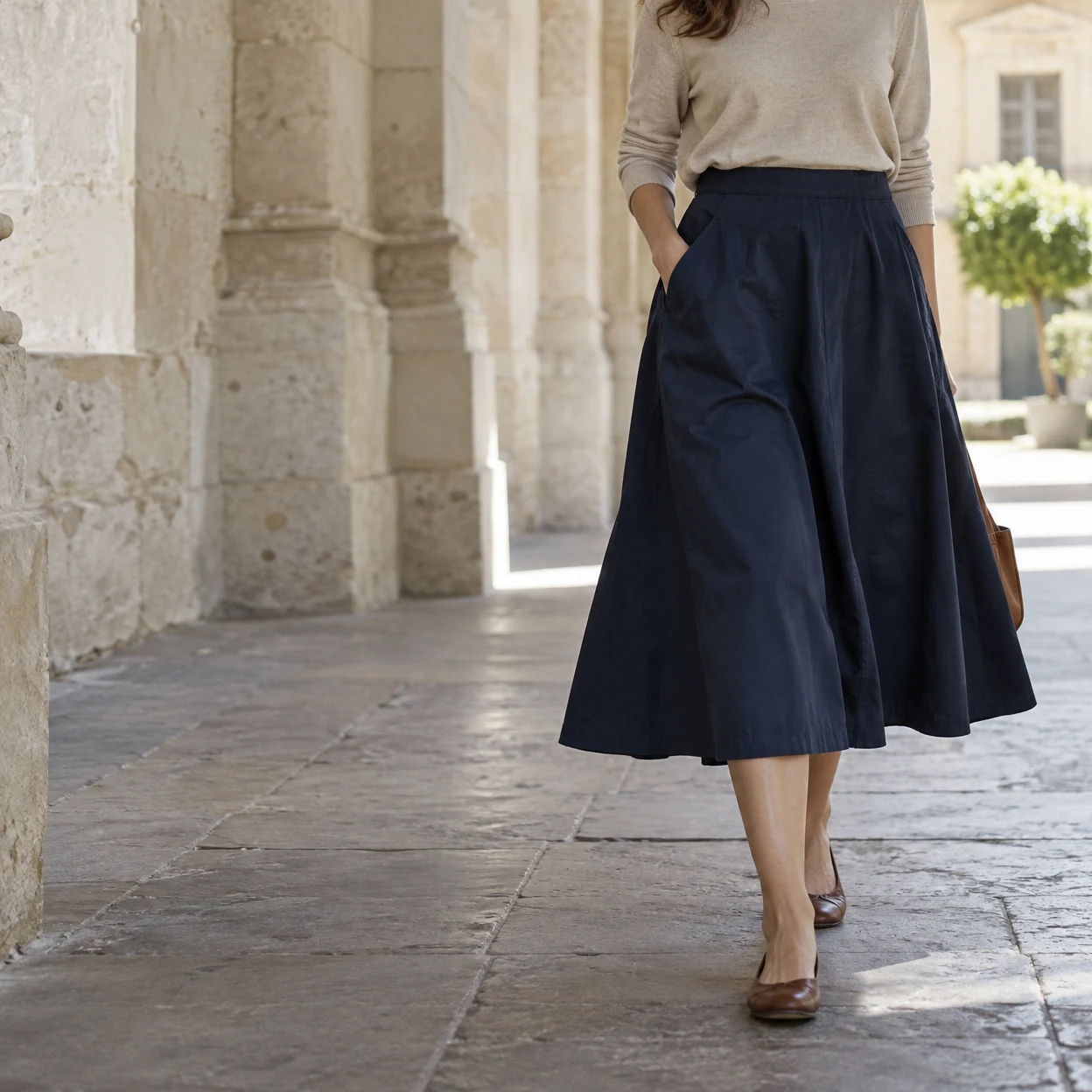 A woman in a navy A-line midi skirt, oatmeal top, and brown flat shoes walks along a shaded limestone arcade, with sunlit paving and a potted tree in the…