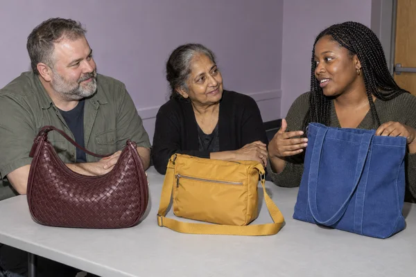Three seated people talk behind a pale grey table displaying an oxblood woven shoulder bag, a mustard yellow zippered crossbody bag, and a blue canvas tote.