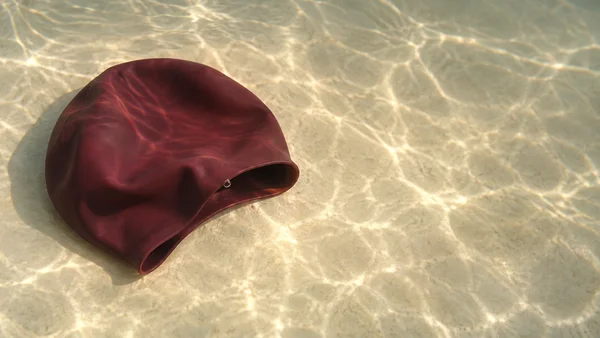 A burgundy swimming cap lies folded on a pale cream underwater tank floor, with its open rim facing right and a small bubble clinging near the opening.
