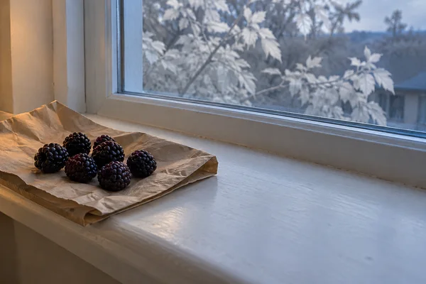 A cluster of seven dark blackberries rests on folded brown paper at the left end of an off-white kitchen windowsill, beside a window with pale leaves visible…