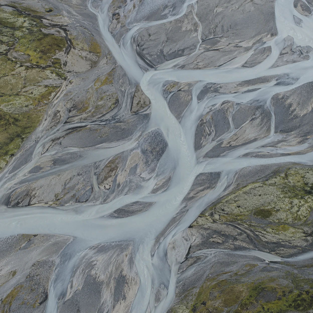 Straight-down aerial view of pale grey braided meltwater channels winding through slate-grey and tan gravel bars, with sparse olive-green patches along the…
