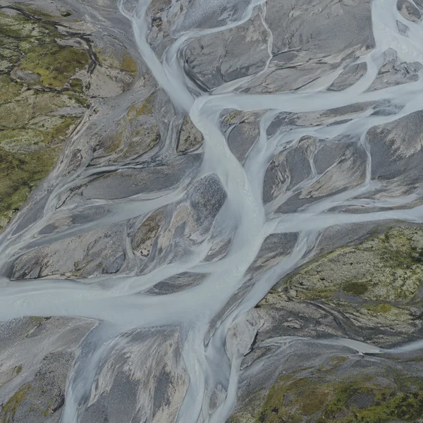 Straight-down aerial view of pale grey braided meltwater channels winding through slate-grey and tan gravel bars, with sparse olive-green patches along the…