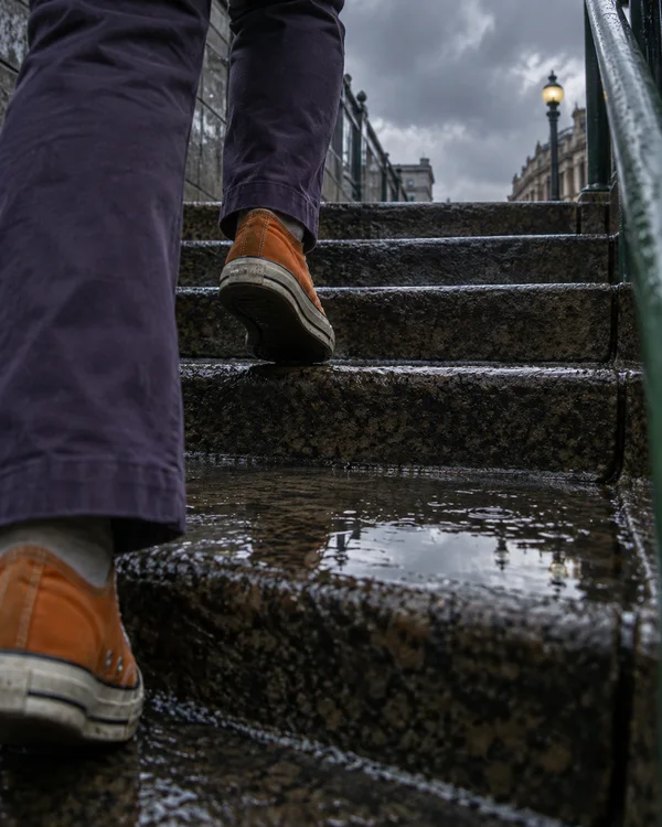 An ankle-height view of a commuter climbing wet stone steps, wearing violet trousers and orange sneakers.